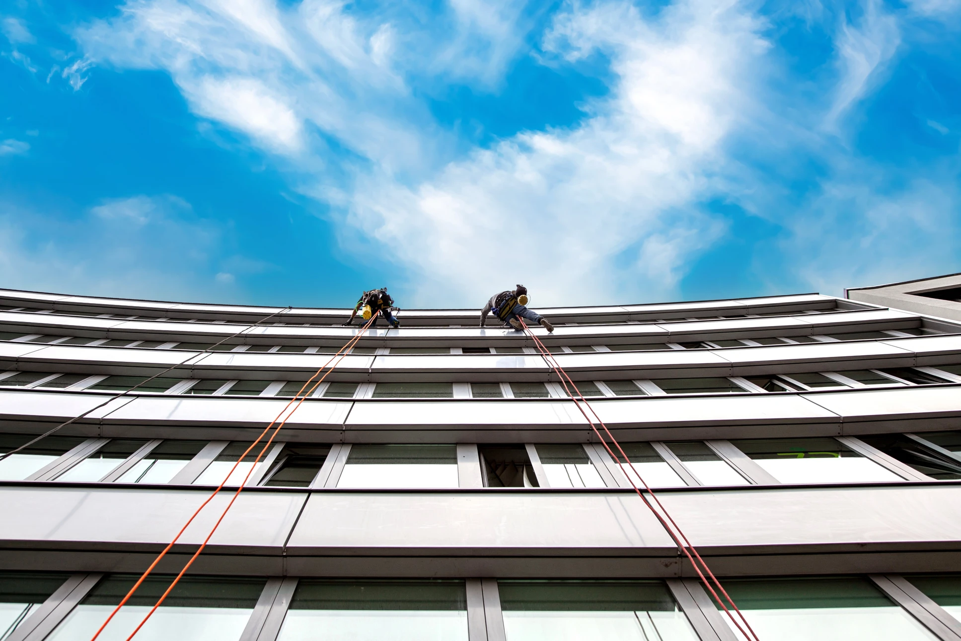 Deux ouvriers équipés de harnais de sécurité effectuent des travaux de nettoyage ou maintenance sur la façade d'un immeuble moderne multi-étages